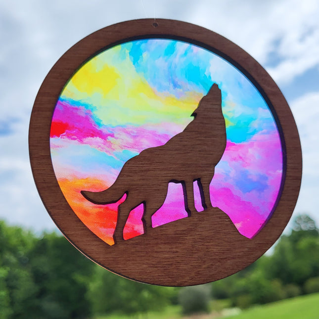 an image of a wooden suncatcher in the shape of a circle. It has a howling wolf on a hill laser cut from the center. The transparent acrylic background shows the "Rainbow" variation with hues of rainbow colors that look "cloud like"