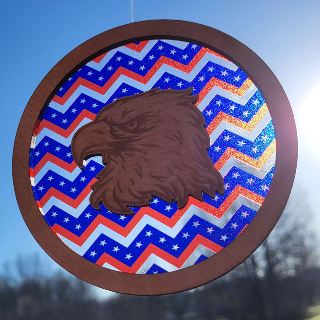 an image of a wooden suncatcher in the shape of a circle. It has an eagle head laser engraved and cut from the center. The transparent acrylic background shows red stripes as well as clear stripes and blue ones with stars