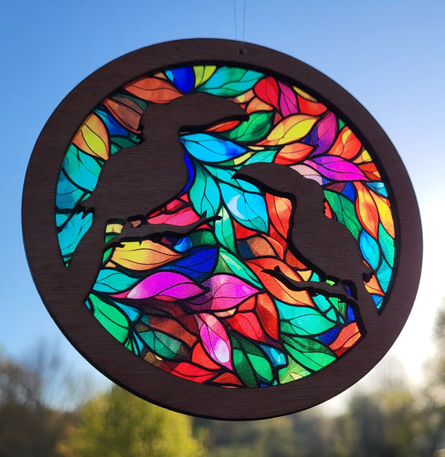 an image of a wooden suncatcher in the shape of a circle. There are 2 toucans laser cut from the top layer. The transparent acrylic background shows a colorful array of leaves