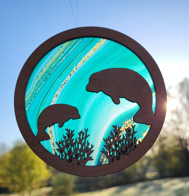 an image of a wooden suncatcher in the shape of a circle. 2 manatees and seaweed are laser cut from the center. The transparent acrylic background has hues of green with random gold wavy lines sometimes coming through the scene