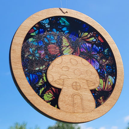 an image of a wooden suncatcher in the shape of a circle with a toadstool/mushroom house laser cut from the middle. The transparent acrylic background shows a multitude of colorful butterflies