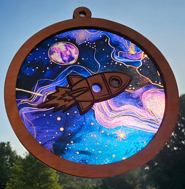an image of a wooden, circular, suncatcher with a rocket ship laser cut in the center. The transparent acrylic background depicts the solar system with blues and purples