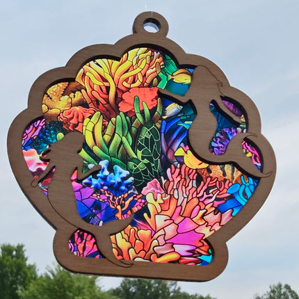 an image of a wooden suncatcher in the shape of a seashell. There are 2 mermaids laser cut from the top layer. The transparent acrylic background shows a colorful reef with fish and coral.