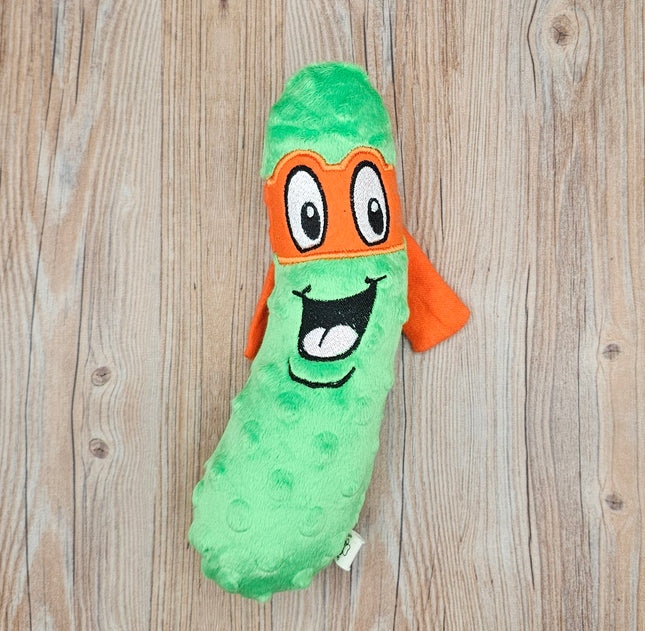 A green plush ninja pickle with an orange eye mask and an embroidered smiling face. The pickle is a plush toy with a textured surface.