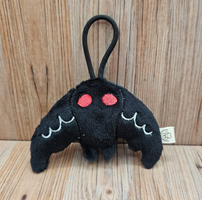 Black plush mothman toy with red eyes and white accents on the wings. Black loop for travel or display from a bag