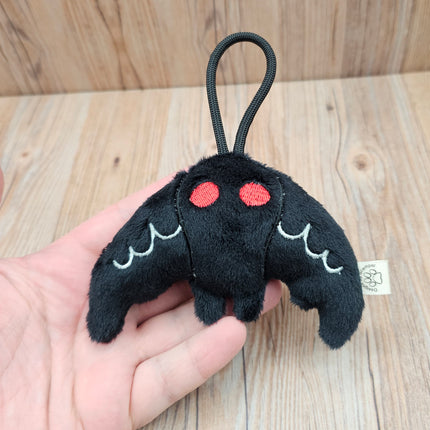 Black plush mothman toy with red eyes and white accents on the wings. Black loop for travel or display from a bag. Held in hand to show size