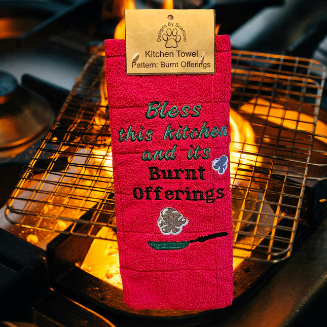image of a red embroidered polyester kitchen towels. On it says "Bless this kitchen and its burnt offerings" and shows a skillet with smoke puffs