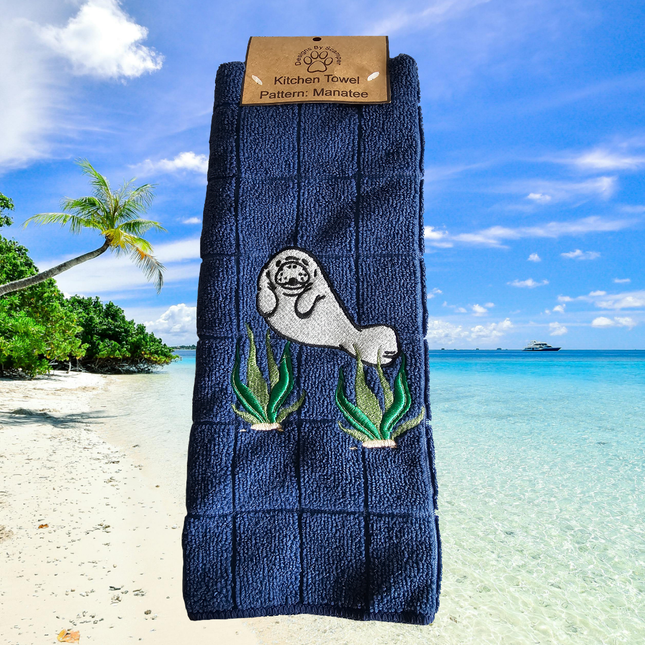 image of a dark blue embroidered polyester kitchen towel. The towel has a cute manatee on the front amongst seaweed