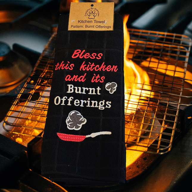 image of a black embroidered polyester kitchen towels. On it says "Bless this kitchen and its burnt offerings" and shows a skillet with smoke puffs