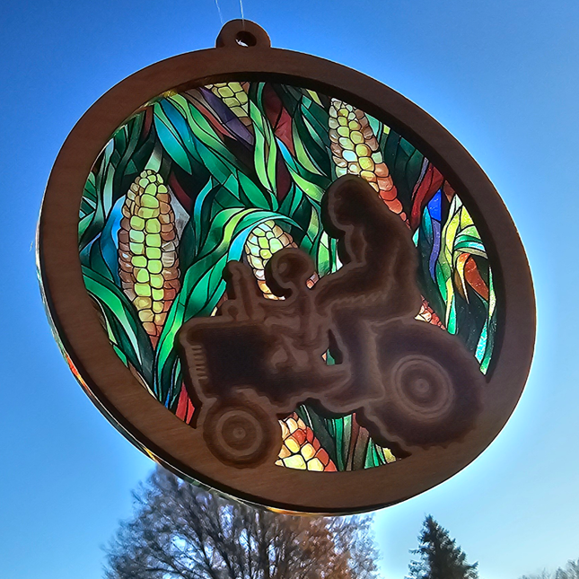 Wooden, circular suncatcher with an engraved alien & Bigfoot riding a tractor. Transparent acrylic background of corn fields.