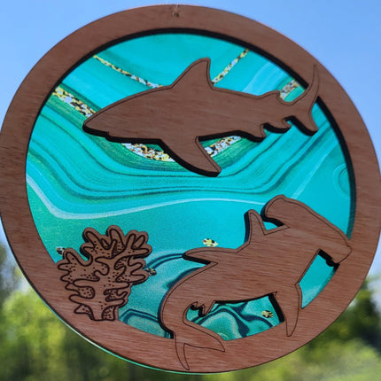 Circular, wooden suncatcher with sharks and seaweed laser engraved and cut in it. The transparent acrylic background has hues of green and flecks of gold.