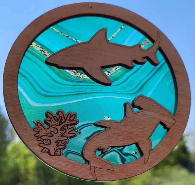 Circular, wooden suncatcher with sharks and seaweed laser engraved and cut in it. The transparent acrylic background has hues of green and flecks of gold.