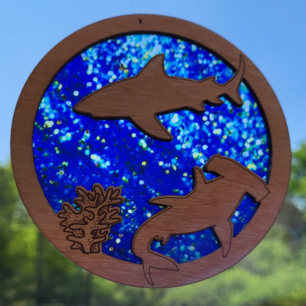 Circular, wooden suncatcher with sharks and seaweed laser engraved and cut in it. The transparent acrylic background has hues of blue and flecks of gold and clear spots that look like bubbles.