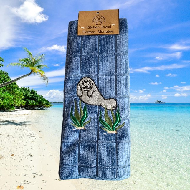 image of a light blue embroidered polyester kitchen towel. The towel has a cute manatee on the front amongst seaweed