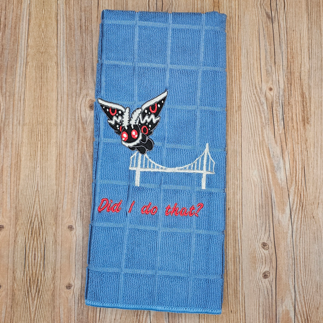 image of a light blue embroidered polyester kitchen towel. It shows mothman flying away from a bridge with the words "did i do that?" underneath