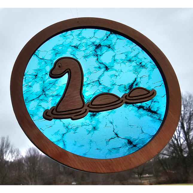 a wooden, circular suncatcher with a cartoon loch ness monster laser etched and cut in the middle. The transparent acrylic background has hues of turquoise and black cracks going through it
