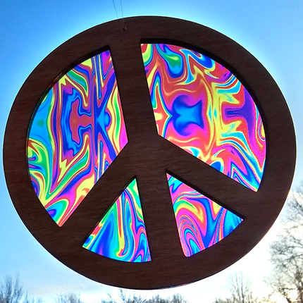 an image of a wooden suncatcher in the shape of a peace sign. The transparent acrylic background is a variety of tie dyed colors