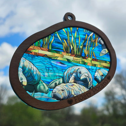 Transparent acrylic "Stained glass" manatee scene in a wooden frame with 'Floaty Potato' laser etched at the bottom. Suncatcher shows variation in placement of manatee scene