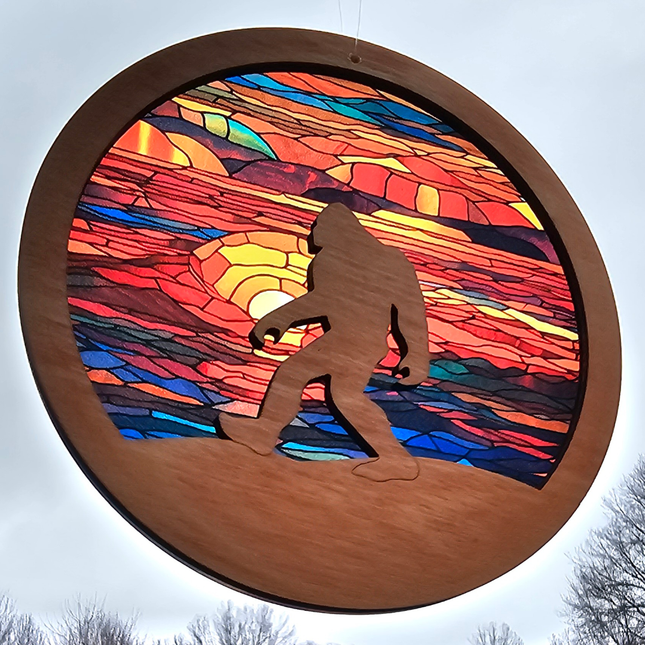 an image of a wooden suncatcher in the shape of a circle with bigfoot laser cut from the center and shown walking over terrain. The transparent acrylic background has a multitude of sunset colors in a stained glass look. 