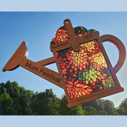 An image of a wooden suncatcher in the shape of a watering can. The transparent acrylic background shows a variety of mums in fall colors. This watering can has the words "Barberton Ohio" and "Mum Festival" laser engraved on it