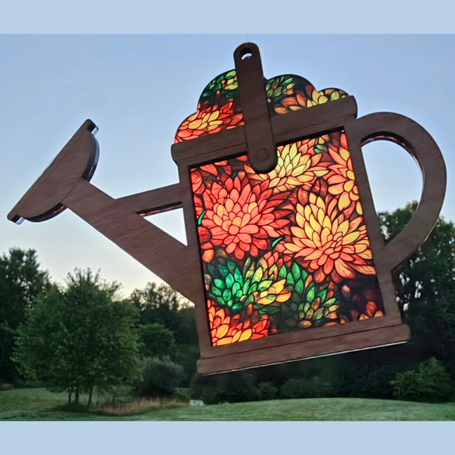 An image of a wooden suncatcher in the shape of a watering can. The transparent acrylic background shows a variety of mums in fall colors